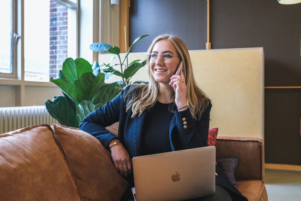 Smiling professional in a navy blazer talking on the phone with a laptop on her lap in a bright office lounge