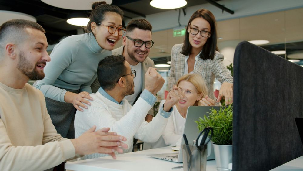 A team of coworkers gathered around a laptop, smiling and cheering together