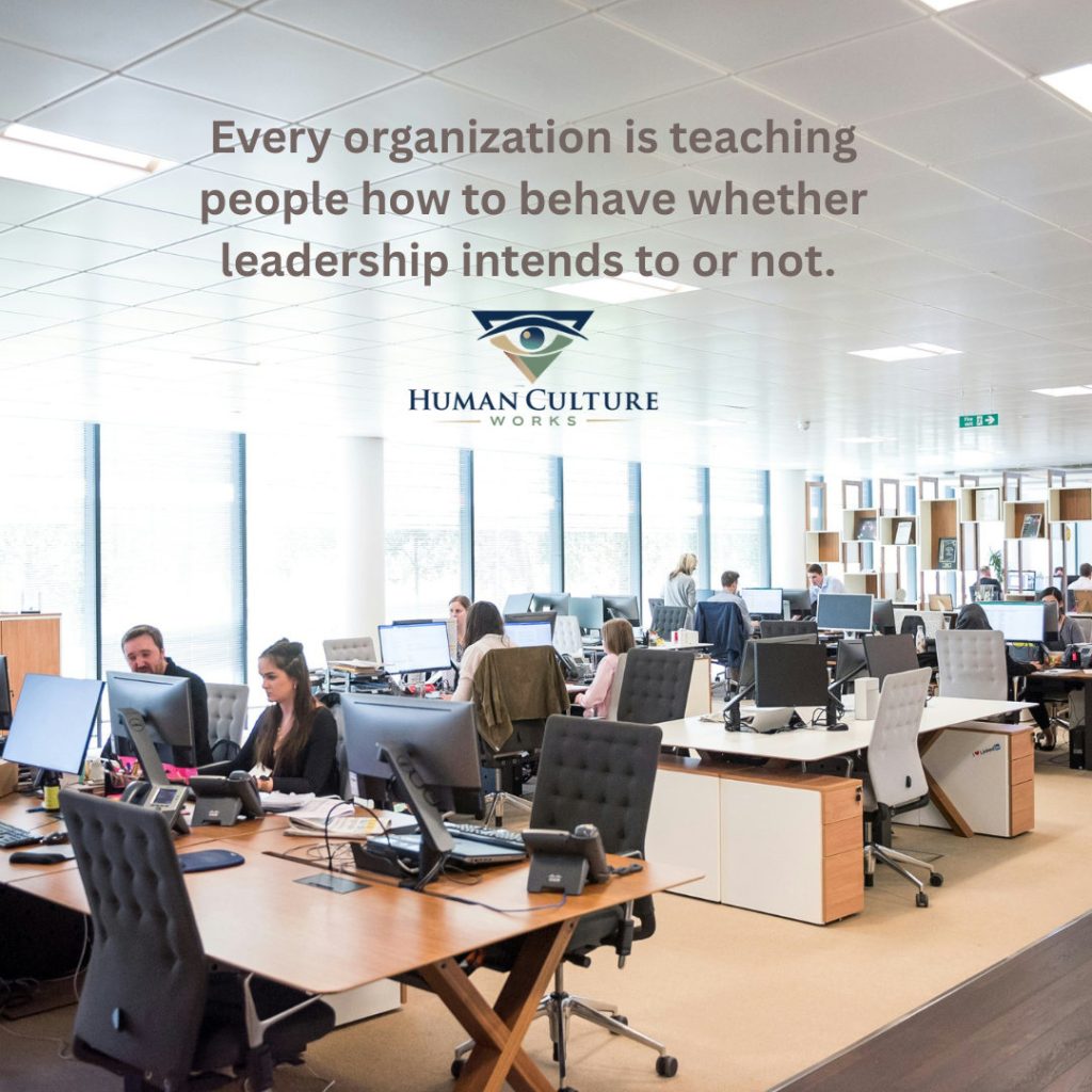 Every organization is teaching people how to behave whether leadership intends to or not, text over an open plan office